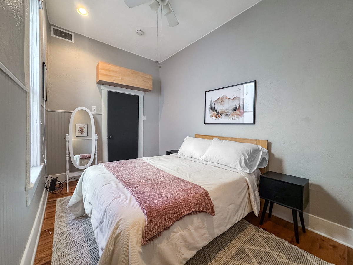 Основната спалня разполага с неутрални произведения на изкуството, ретро огледало в цял ръст и две нощни лампи (не са показани).