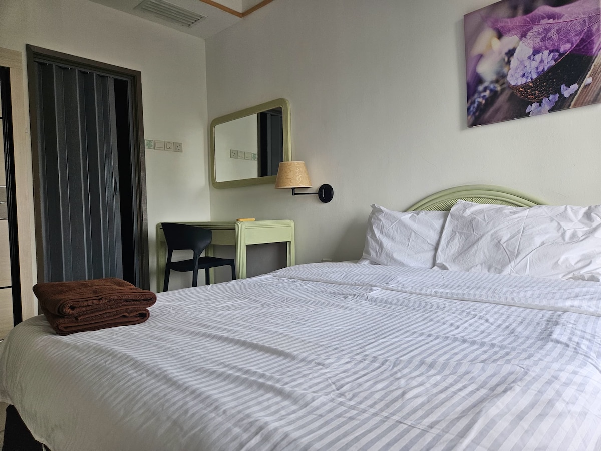 A spacious sleeping area is framed by light-colored walls and features a striped bedding arrangement. A neatly folded set of towels is placed on the bed. A round mirror above a green desk reflects the room's soft lighting, while a wall-mounted lamp provides additional illumination.