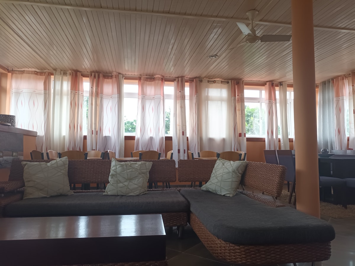 A spacious seating area is highlighted by a large, comfortable sofa with soft cushions. Tall windows dressed in light fabric allow natural light to fill the room, complemented by potted plants that provide a touch of greenery.