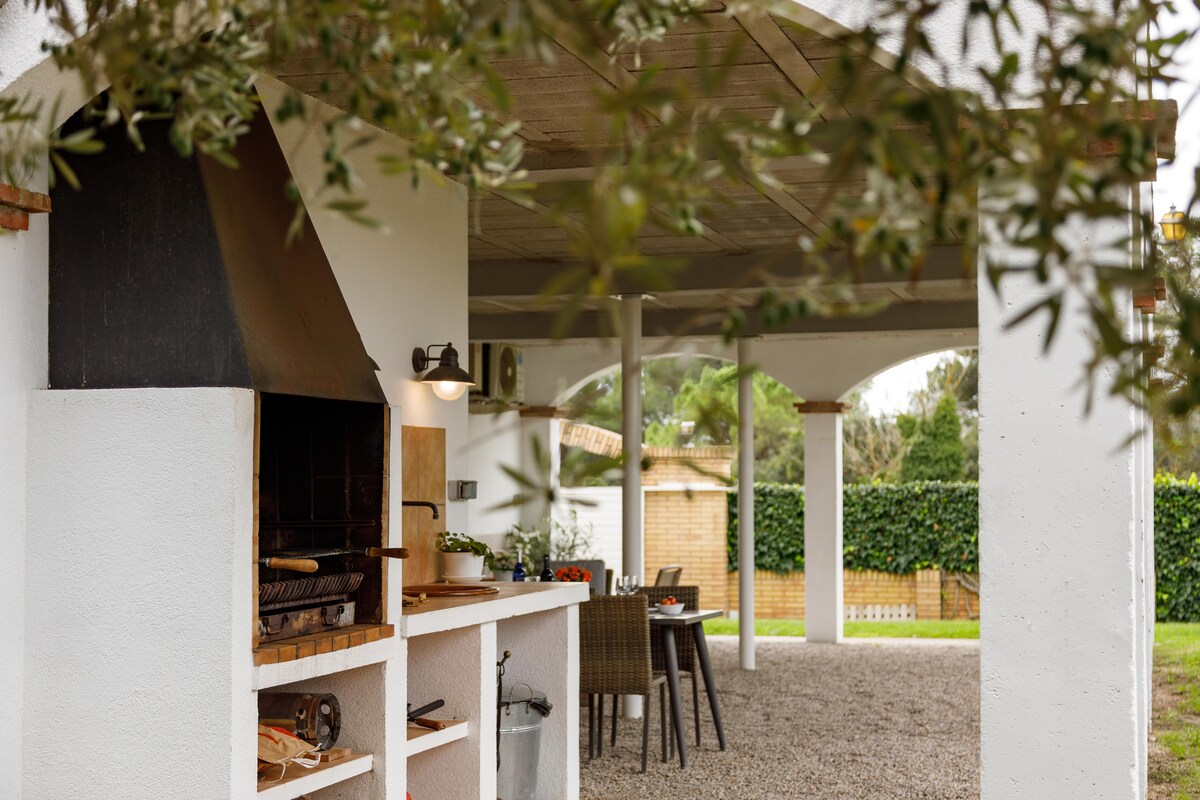An outdoor barbecue area is visible, featuring a built-in grill adjacent to a countertop. A set of dining chairs is arranged nearby under a covered terrace, providing shaded seating options. Lush greenery surrounds the space, creating a relaxed atmosphere for outdoor meals.