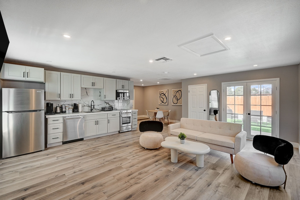 A spacious living area features modern cabinetry and stainless steel appliances in the kitchen, with a dining table for four. A plush sofa and circular lounge chairs invite relaxation, while natural light flows through large doors that open to the fenced patio.