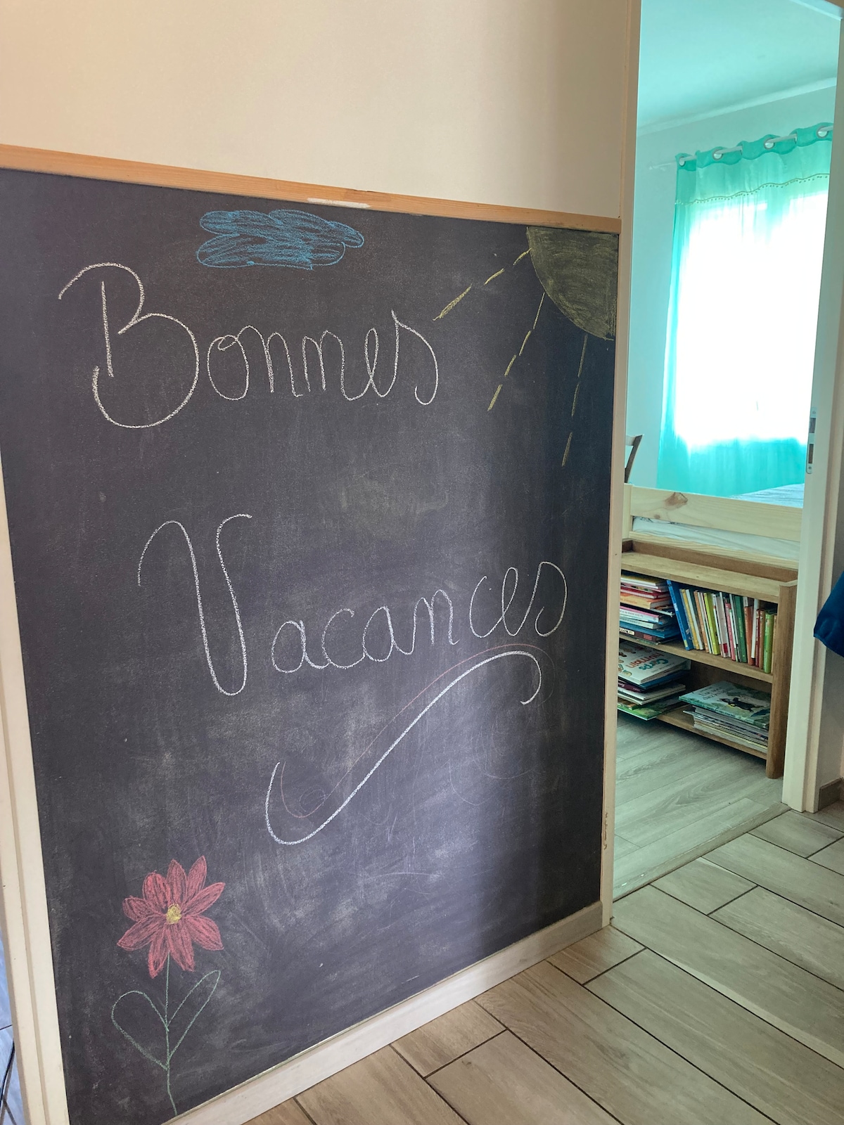 A chalkboard welcomes guests with the message 'Bonnes Vacances,' complemented by colorful drawings of flowers and the sun. A bright room with turquoise curtains and a bookshelf filled with books is visible in the background, adding to the inviting atmosphere.