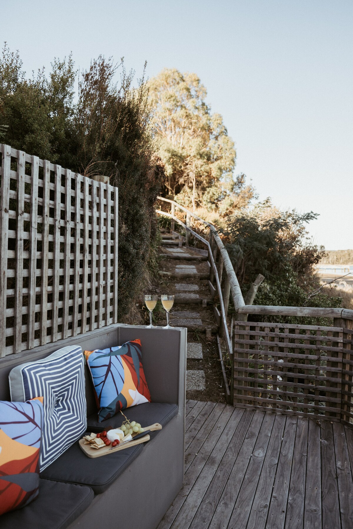 A comfortable outdoor seating area is shown, featuring colorful cushions atop a grey sofa. Two glasses of champagne are placed on a small wooden table, inviting relaxation. A winding pathway lined with greenery leads upward, blending harmoniously with the natural surroundings.