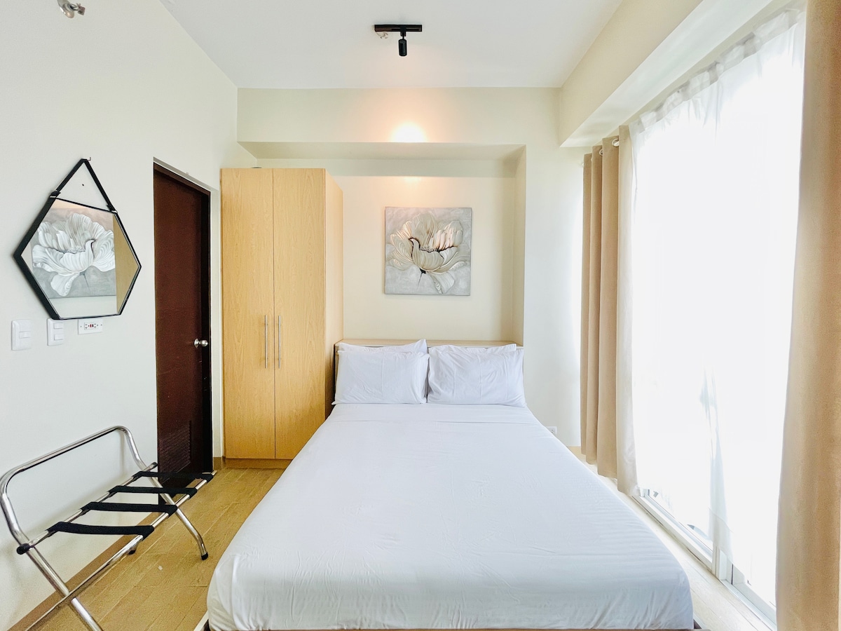 A bright sleeping area features a spacious bed with crisp white linens. A large window is draped with light-colored curtains, allowing natural light to fill the room. A simple piece of art hangs above the bed, and a wardrobe is positioned beside a door.
