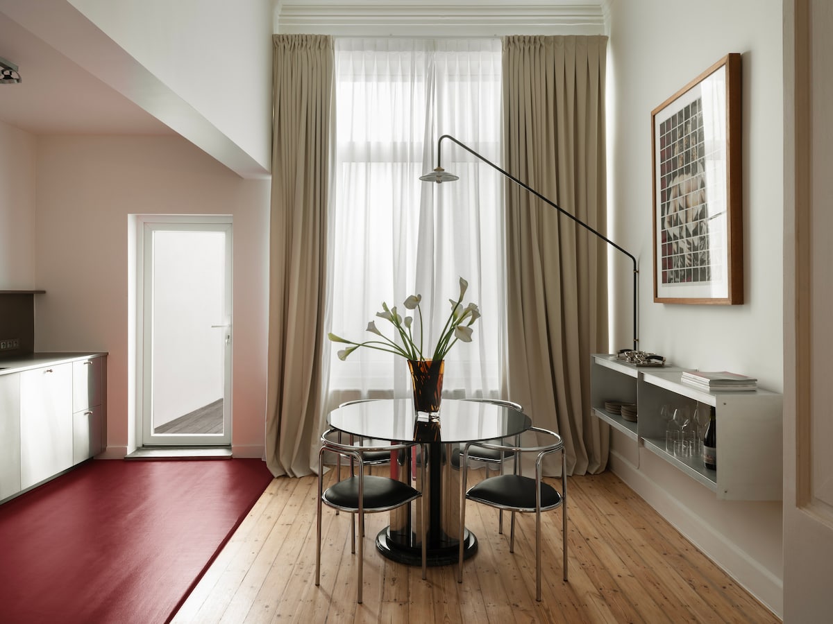 A dining area features a round table surrounded by four black chairs. A vase with white flowers sits at the center. Natural light is diffused through large curtains, and a door provides access to an outdoor space. A wall-mounted shelf displays glassware and a record player.