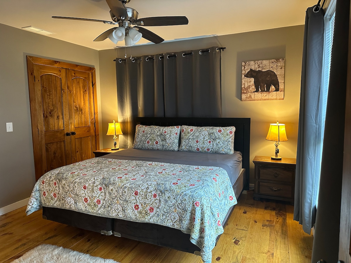 A king bedroom is presented, featuring a dark wood bedframe with a floral-patterned comforter. Two bedside tables with lamps provide illumination, and a bear art piece is mounted on the wall. Light flows in from a window draped with dark curtains, enhancing the room's cozy feel.
