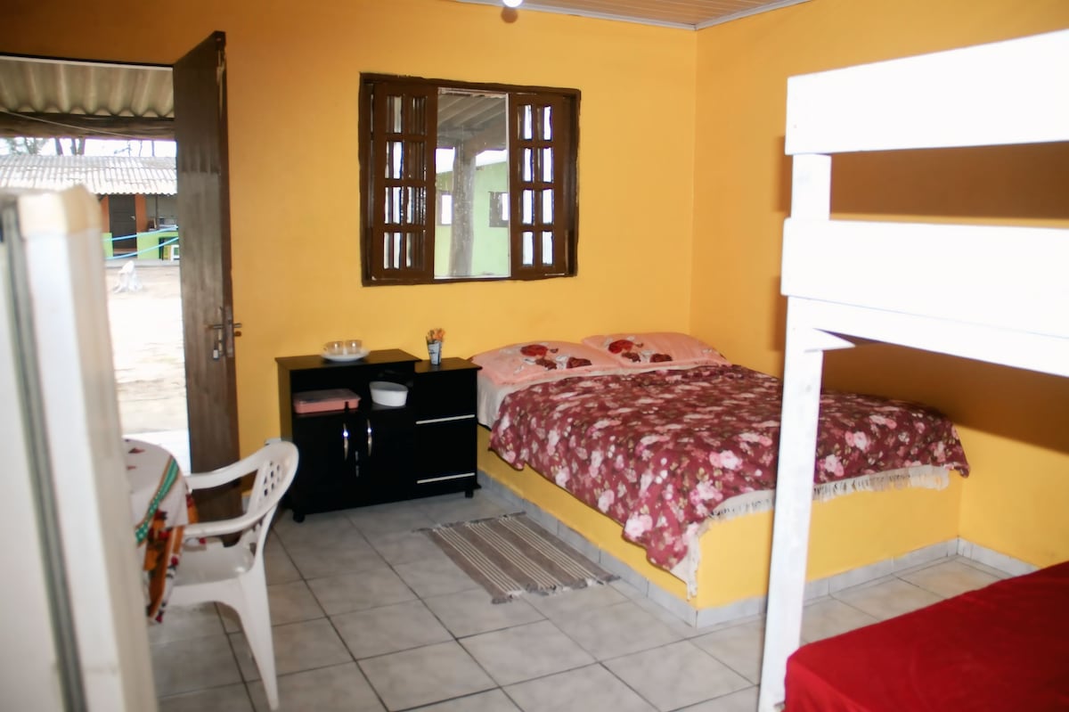 A room features a spacious bed with floral bedding and a bunk bed against a vibrant yellow wall. A dresser holds essentials, and a small table and chair are positioned nearby. Light enters through windows with wooden shutters, enhancing a cozy atmosphere.
