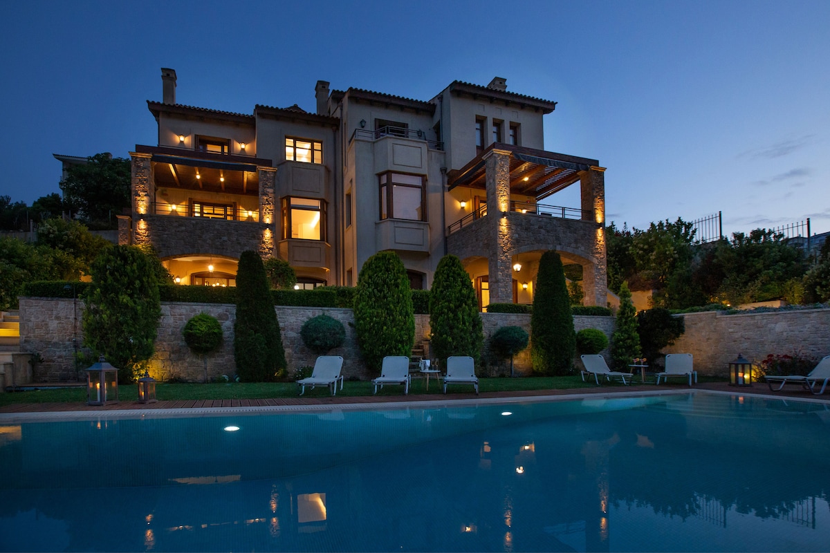 The villa is showcased at dusk, featuring elegant architectural details and well-lit balconies. The inviting swimming pool reflects the soft glow of lights surrounding the property, framed by neatly manicured greenery and lounge chairs arranged around the pool area.