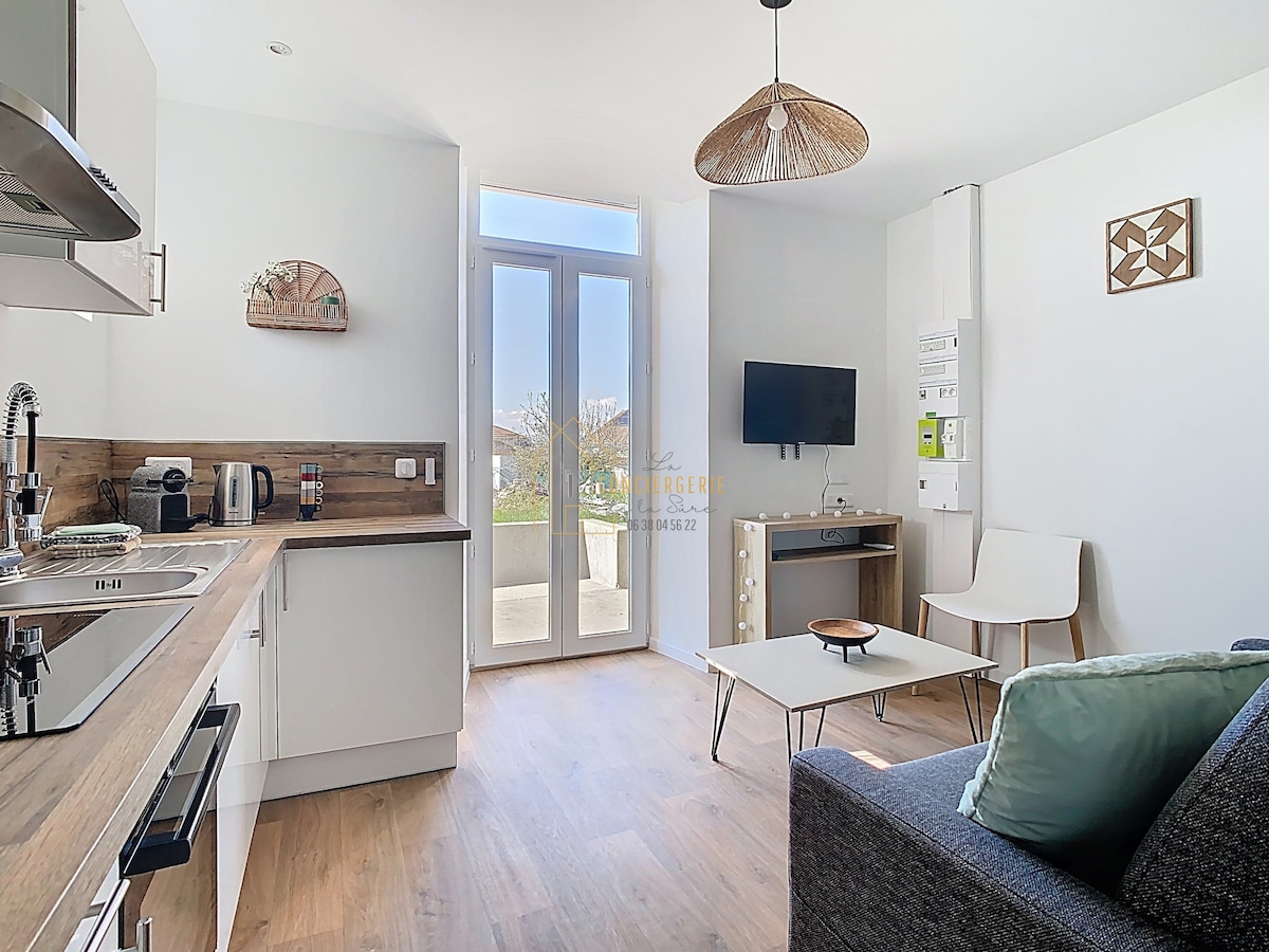 The image displays a bright studio with modern furnishings. A comfortable gray sofa is positioned near a light wood coffee table. A kitchenette is visible with stainless steel appliances and wooden countertops. Large glass doors allow natural light and open to a view of the garden.