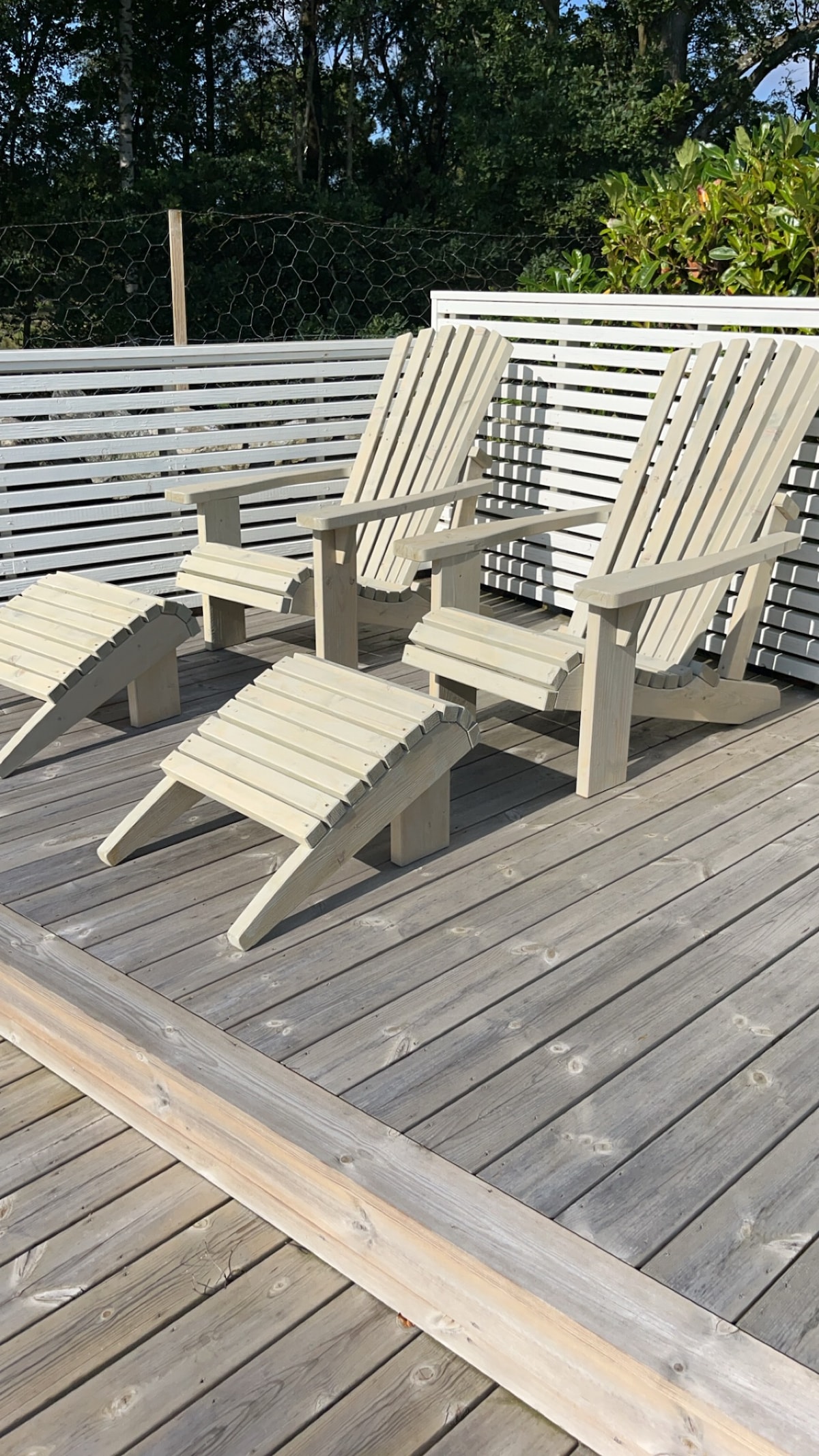 Two light-colored Adirondack chairs are positioned on a wooden deck, accompanied by matching footrests. A wooden railing surrounds the area, offering a sense of privacy. The setting is partially shaded by lush greenery in the background, providing a relaxed outdoor space.