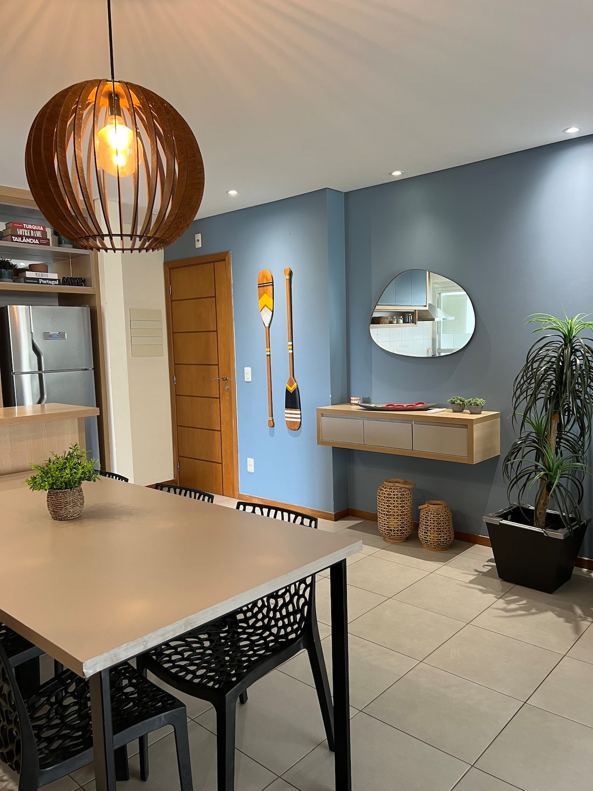 A modern dining area features a rectangular table surrounded by six black chairs. A wooden pendant light hangs above the table. A mirror is mounted on a blue wall, accented by decorative paddles and a small indoor plant on a side table.