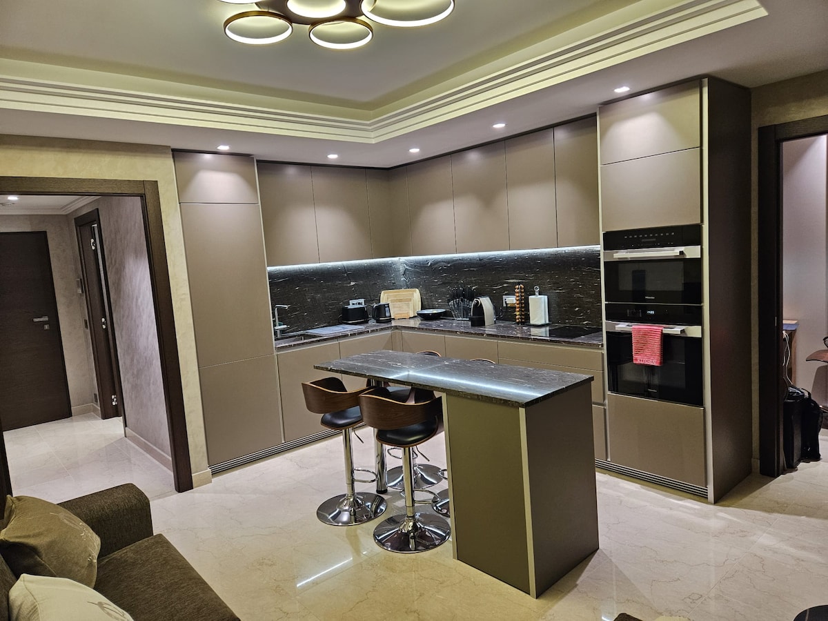 A modern kitchen features sleek cabinetry and a granite countertop, illuminated by stylish overhead lighting. High-backed stools are positioned at the island, and appliances are integrated into the cabinetry, blending functionality with contemporary design.