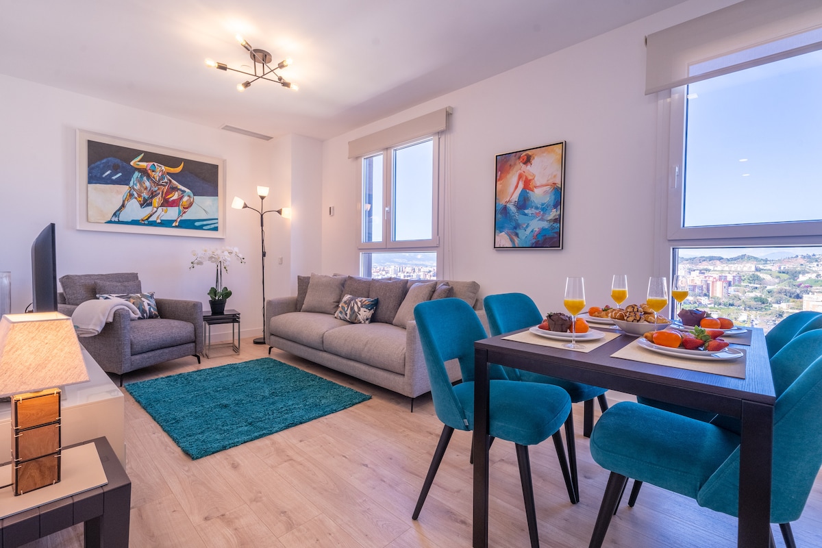A spacious living area features a comfortable grey sofa, two accent chairs in vibrant teal, and a dining table set for two with a selection of fruits and beverages. Large windows provide ample natural light and impressive city views, complemented by modern artwork and elegant lighting fixtures.