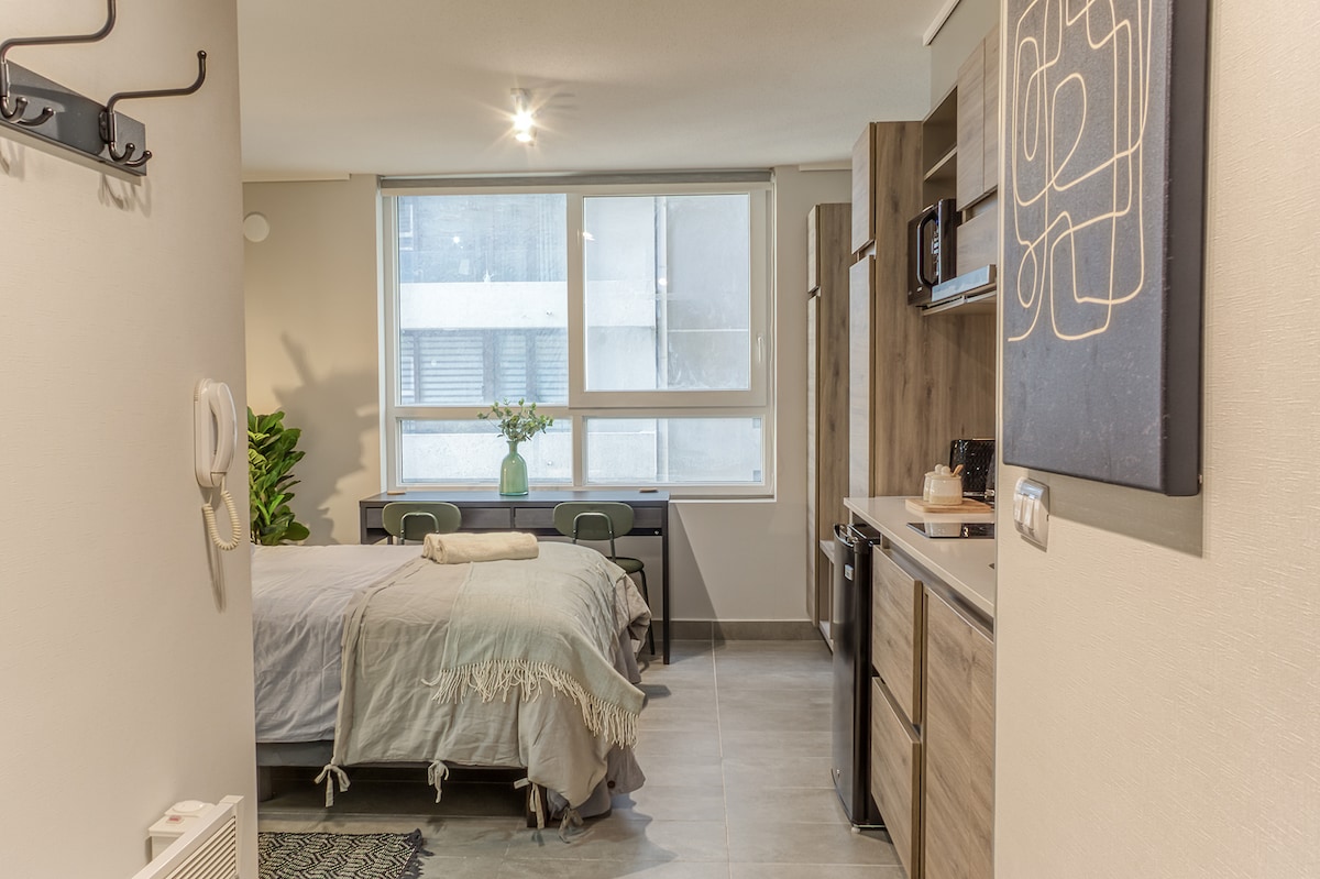 A cozy studio space is highlighted by a spacious bed draped in a light-colored blanket. A functional kitchenette and dining area can be seen, accompanied by a window allowing natural light to fill the room. Decorative elements, including a wall art piece and potted plants, enhance the ambiance.