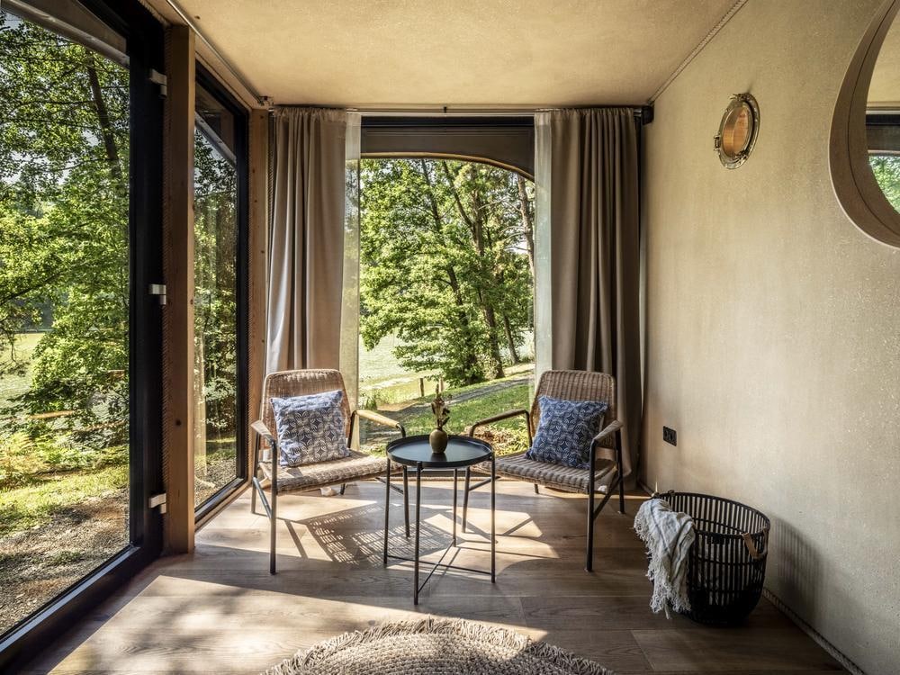 A cozy seating area is presented, featuring two woven chairs and a round table, positioned near large windows that frame views of the surrounding greenery. Soft curtains filter natural light, while a decorative mirror reflects the serene atmosphere.
