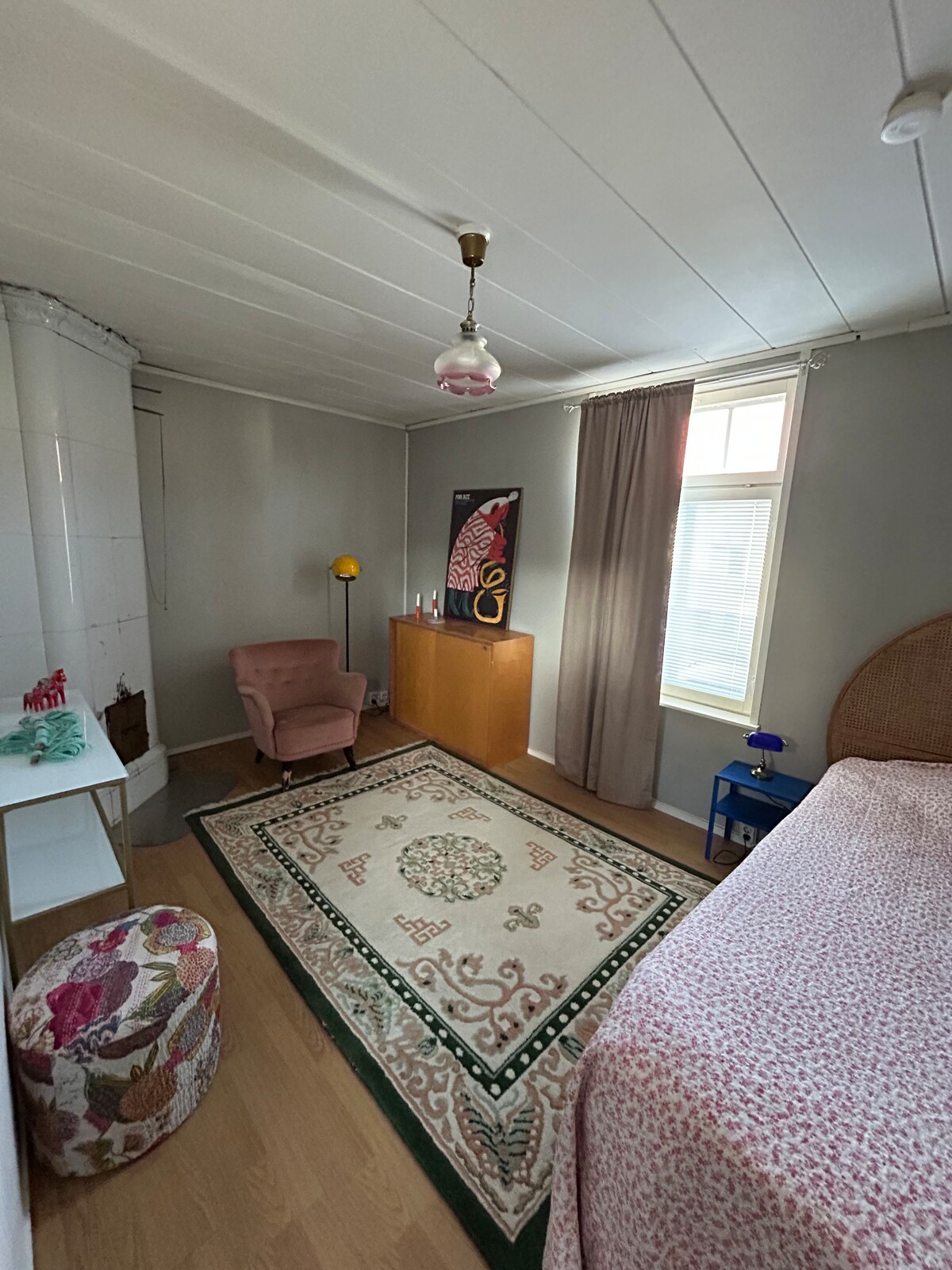 A spacious bedroom is shown with a cozy pink chair beside a storage unit. A rug with intricate patterns covers the floor, complementing the light walls. Natural light enters through a window dressed with light curtains, highlighting the floral details in the decor.