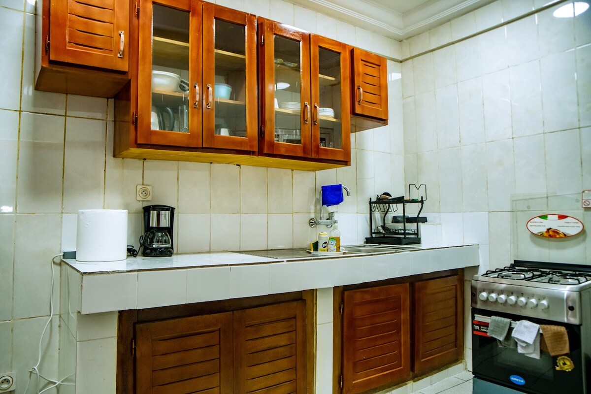 A functional kitchen space features wooden cabinets with glass doors, displaying various dishes and kitchenware. A stainless steel sink is positioned below a window. The counter is equipped with an electric coffee maker, and a gas stove is positioned against the wall.