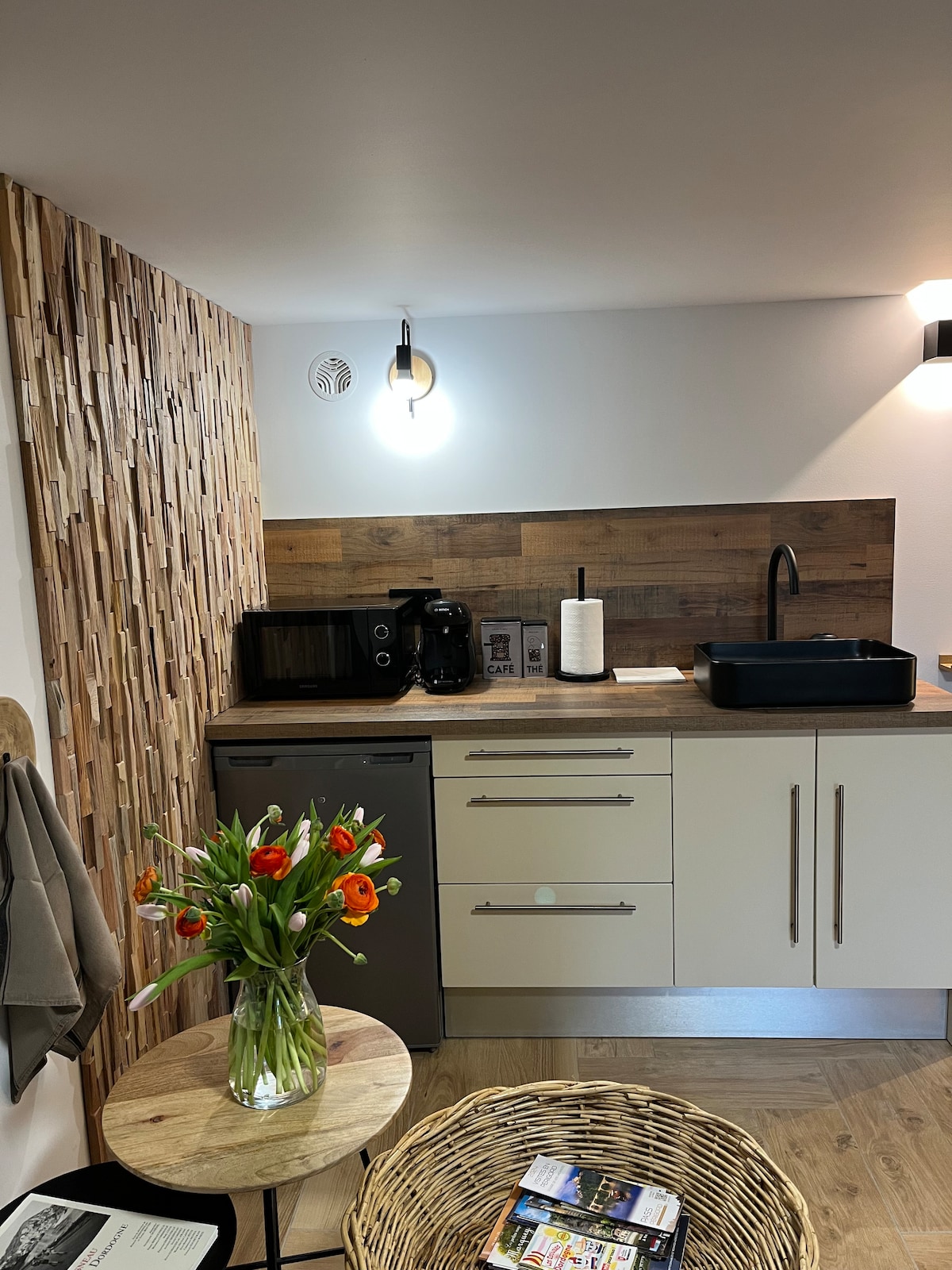 The kitchenette features modern appliances, including a refrigerator, microwave, and coffee maker. A decorative wooden wall adds texture, while a round table holds a vase of vibrant flowers. Natural light enhances the space, creating a functional and inviting area for meal preparation.