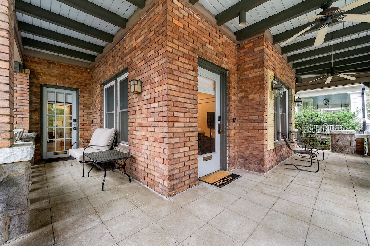 A covered front porch is depicted, featuring brick walls and a tiled floor. Two ceiling fans are visible for comfort. A small table with chairs and a lounge chair are positioned to create a welcoming seating area. Natural light filters in through the adjacent glass doors.