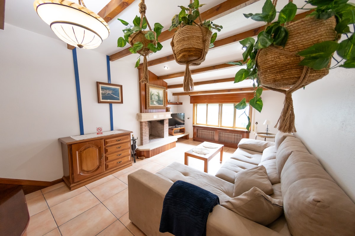 A spacious living room is illuminated by natural light filtering through large windows. A cream-colored sectional sofa is positioned around a wooden coffee table. Green plants hang from the ceiling, complementing a traditional fireplace and wooden storage unit against the wall.