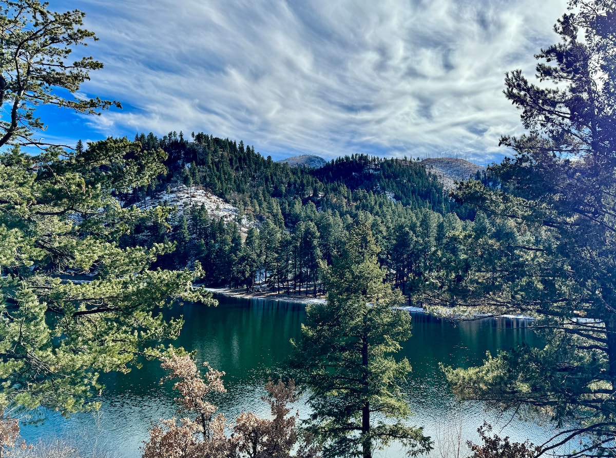 A serene view of a lake is captured, surrounded by lush green trees and distant mountains. The calm water reflects the sky, which is filled with soft clouds. The scene conveys a sense of tranquility and natural beauty.