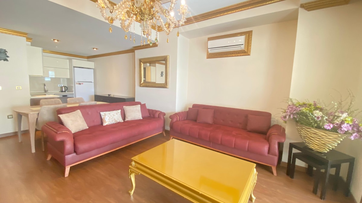 A bright living area features two red sofas arranged around a gold coffee table. A chandelier hangs overhead, providing illumination. A wall mirror and a large vase of flowers add visual interest, while a glimpse of the kitchen is seen in the background.