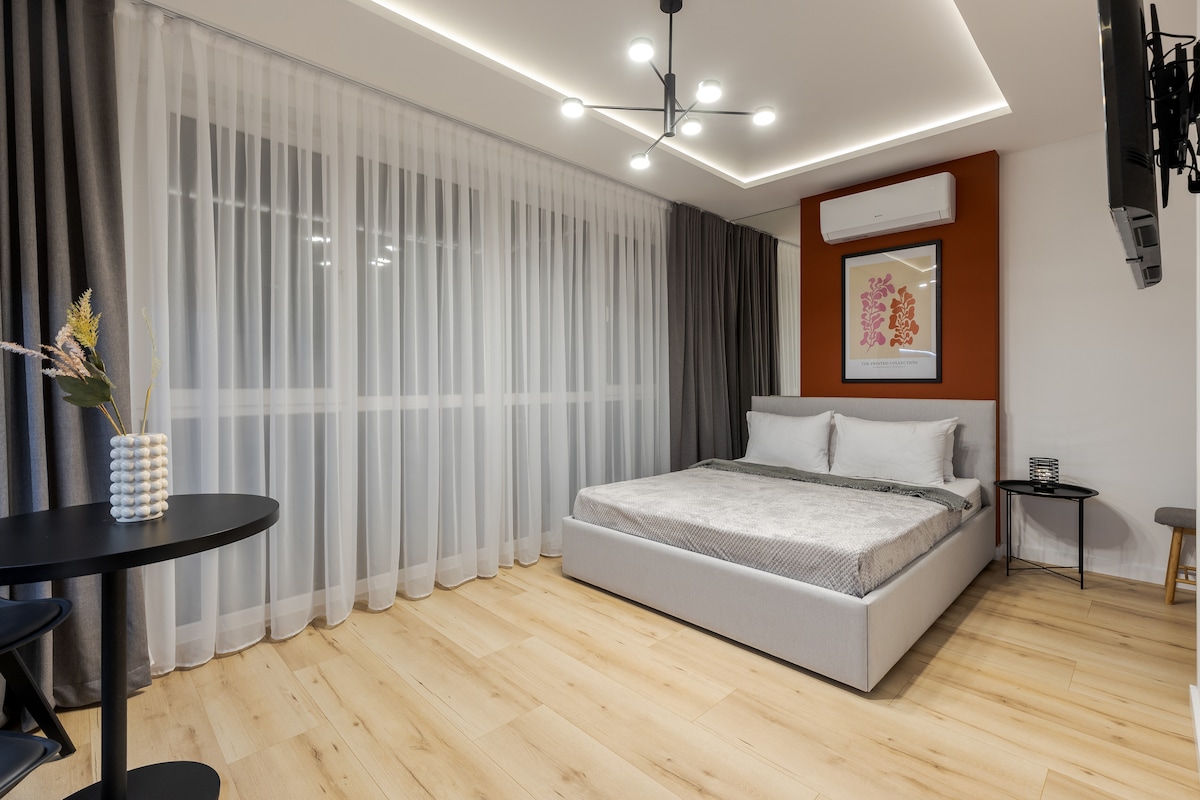 A spacious studio is showcased with light wood flooring and large windows covered by sheer curtains, allowing natural light to fill the room. The sleeping area includes a large bed with soft bedding, while a small dining table is positioned nearby, enhancing the open layout.