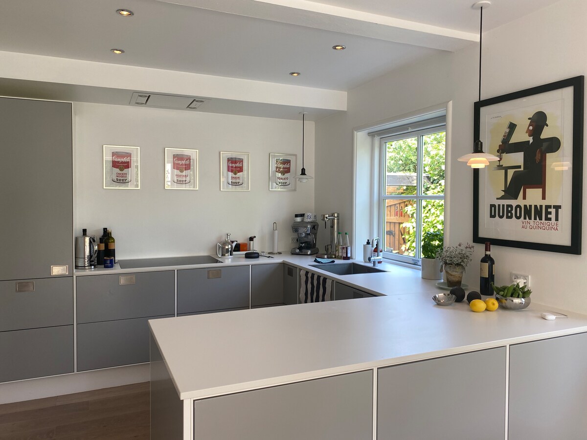 The kitchen features a modern design with sleek gray cabinetry and a spacious island. Natural light streams in from a large window, illuminating various kitchen appliances and decorative items. Framed artworks adorn the walls, adding character to the inviting cooking space.