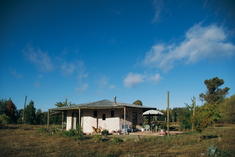 A charming octagonal house sits against a clear blue sky, featuring an earthy exterior with stone accents. A covered porch area provides seating and shade, surrounded by lush greenery and natural landscaping, creating a serene environment that invites relaxation.