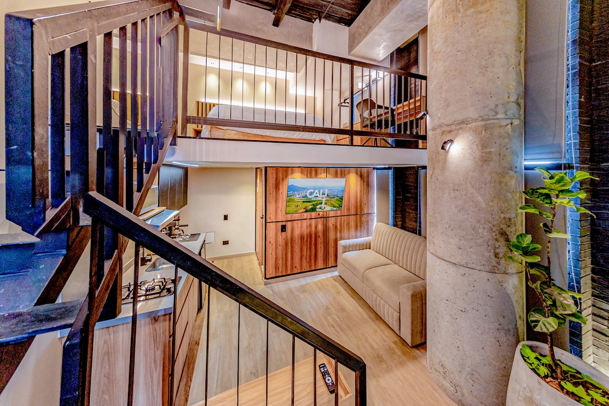 A modern duplex layout showcases a spacious living area featuring a sofa bed and a kitchen with sleek cabinetry. The mezzanine level is visible above, housing the sleeping area. Industrial design elements, including concrete pillars, enhance the ambiance, while natural light flows throughout the space.