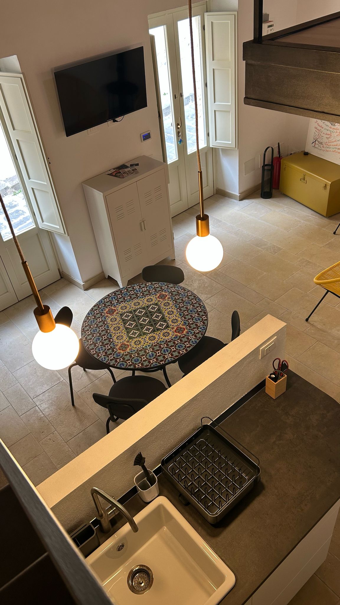 An overhead view reveals a modern kitchen and dining area with a round patterned table surrounded by four chairs. Two pendant lights hang above the table. A television is mounted on the wall, and large windows allow natural light to fill the space.