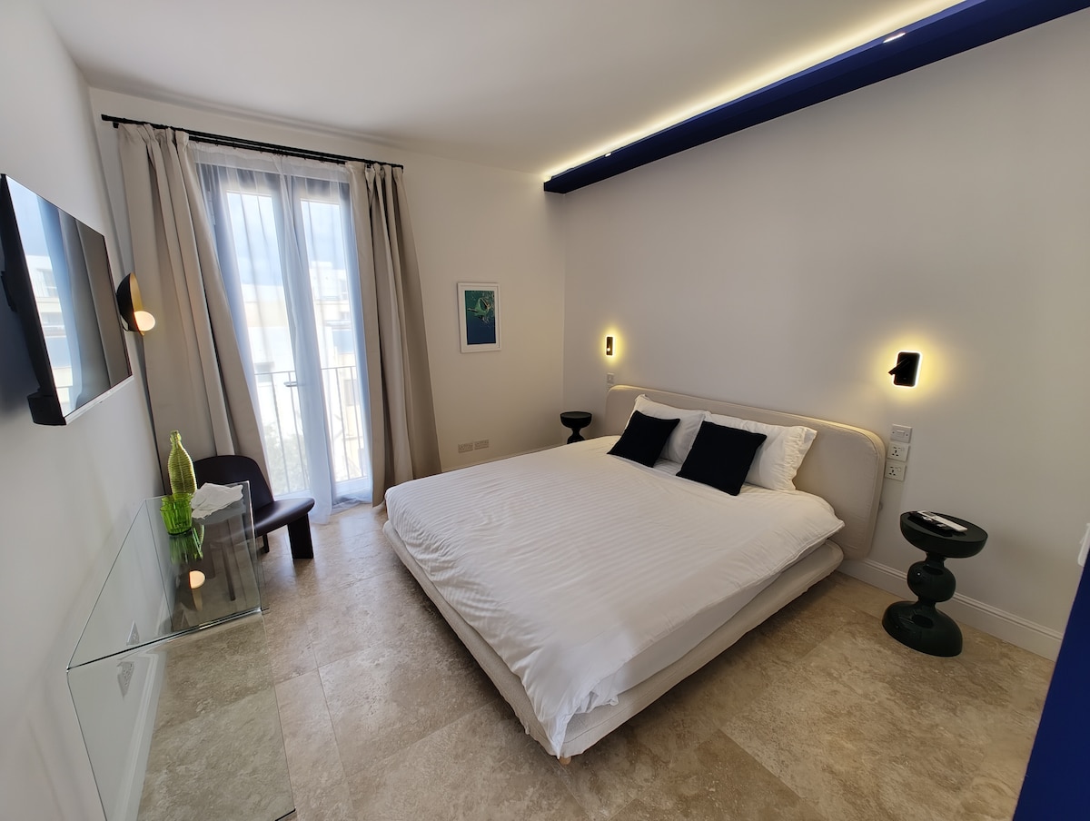 A spacious room is presented with a large bed featuring crisp white linens. Two decorative pillows in navy add contrast. A glass desk with a green vase is visible, accompanied by a modern chair. Natural light filters through sheer curtains, creating a bright environment.