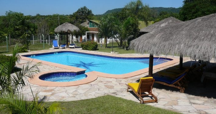 A tranquil outdoor space features a clear blue swimming pool surrounded by a stone patio. Thatched-roof cabanas provide shaded seating, while lounge chairs are positioned near the pool. Lush greenery and distant hills are seen in the background, enhancing the natural setting.