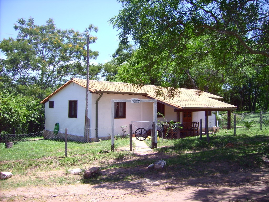 A single-story cottage is framed by greenery and dotted with trees. The building features a sloped roof with tiles and a welcoming porch adorned with a rustic wooden table and chairs. A fence outlines the property, enhancing the secluded natural setting.