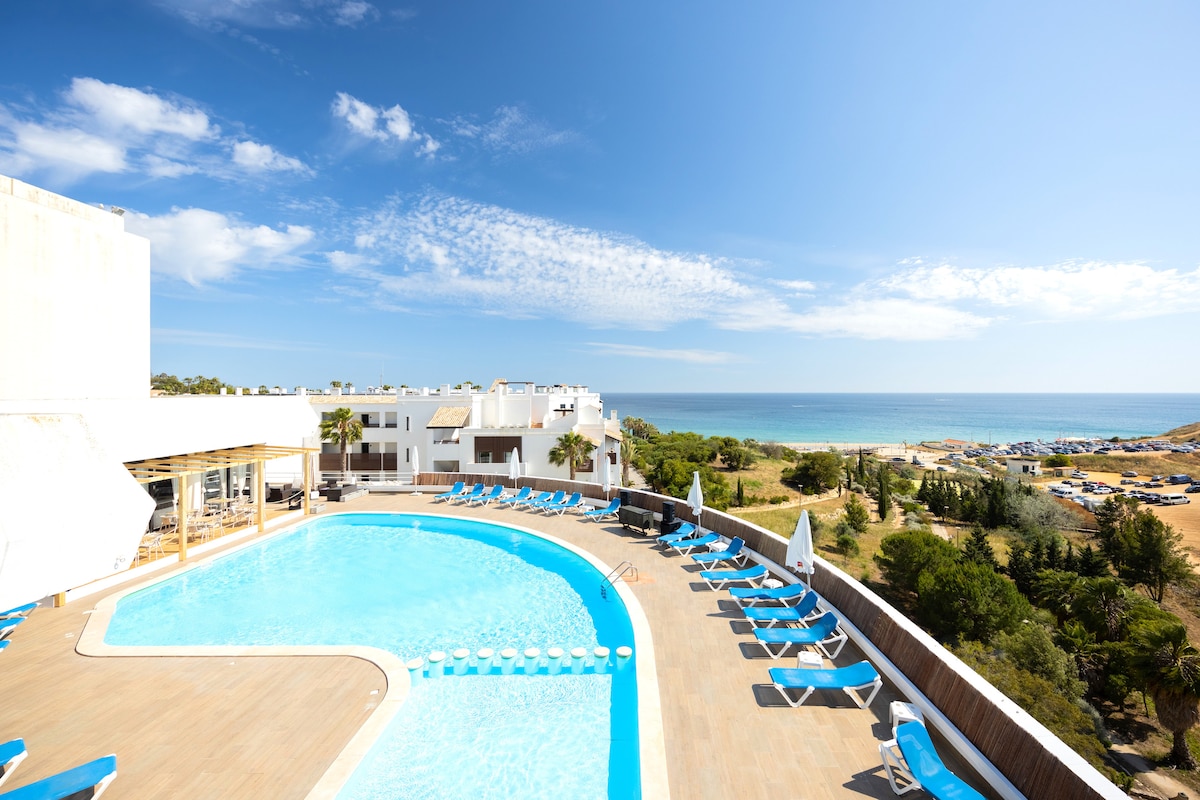 A swimming pool with a curved design is set against a backdrop of the ocean. Lounge chairs are arranged around the pool, and greenery is visible in the distance, along with beach access. The clear blue sky enhances the tranquil atmosphere of the area.