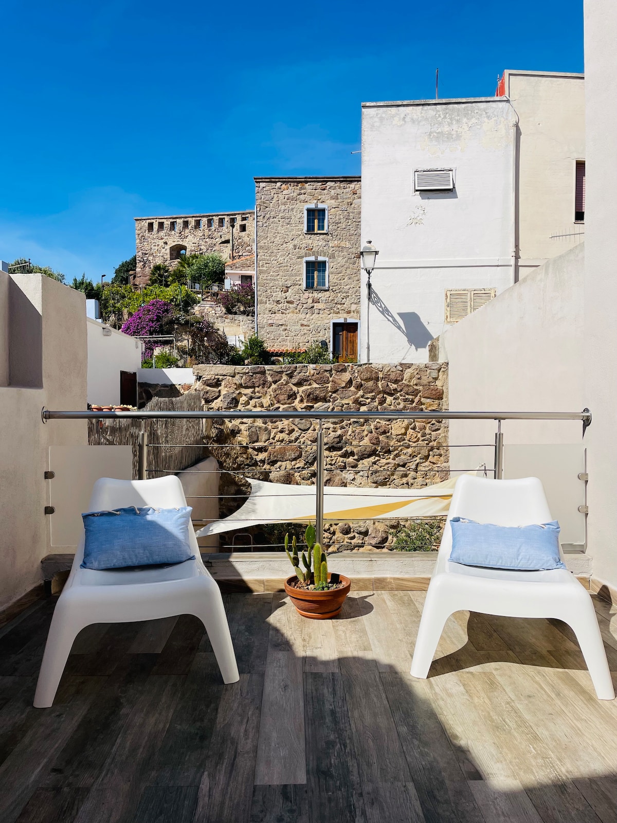 A small outdoor terrace is presented with two white chairs, each topped with a light blue cushion. A potted plant rests on the floor, while the backdrop features a stone wall and historic buildings under a clear blue sky.
