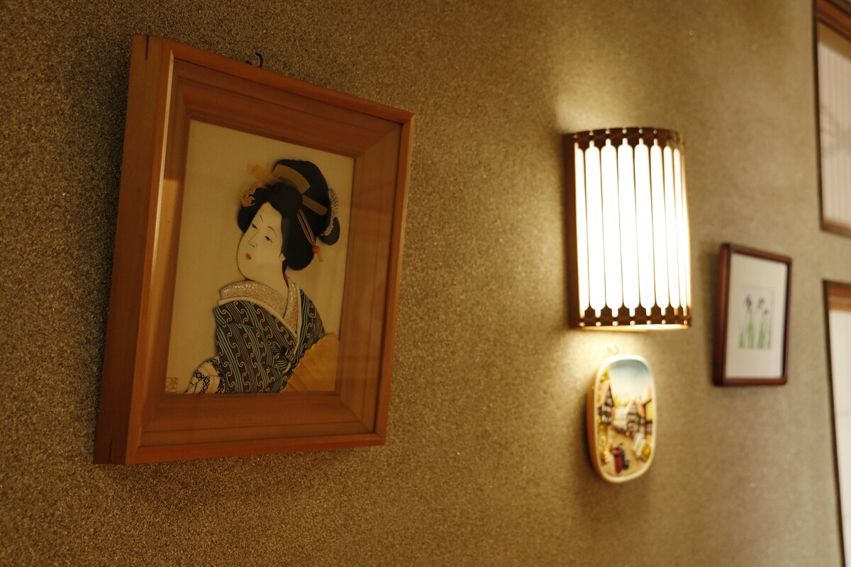 A traditional-style wall showcases a framed artwork depicting a figure in a kimono. A softly glowing wall sconce complements the art, casting gentle light on the textured surface behind. Additional framed images and decorative plates can be seen on the wall, enhancing the cultural ambiance.