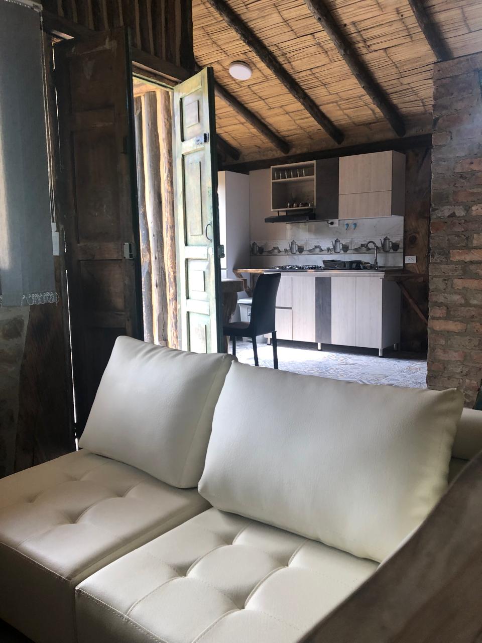 A light-colored sofa is positioned in the foreground, featuring soft cushions. In the background, an open door reveals a kitchen area complete with wooden cabinets and various kitchen utensils. The rustic interior showcases a combination of natural materials, with walls made of brick and a thatched ceiling.