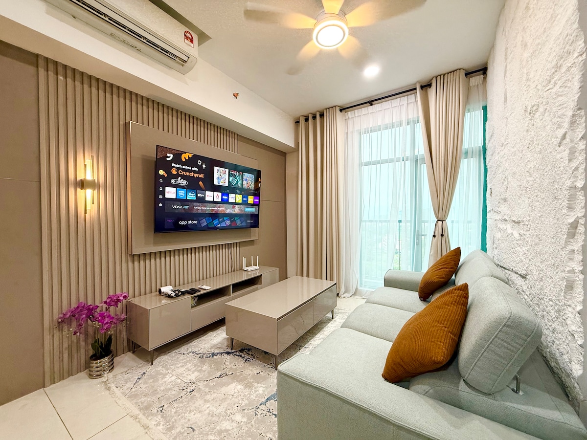 A comfortable living area is presented, featuring a light grey sectional sofa adorned with two mustard-colored cushions. A flat-screen TV is mounted on the wall, displaying various channels. Natural light enters through large windows, with soft curtains framing the view. Decorative plants add subtle touches to the space.