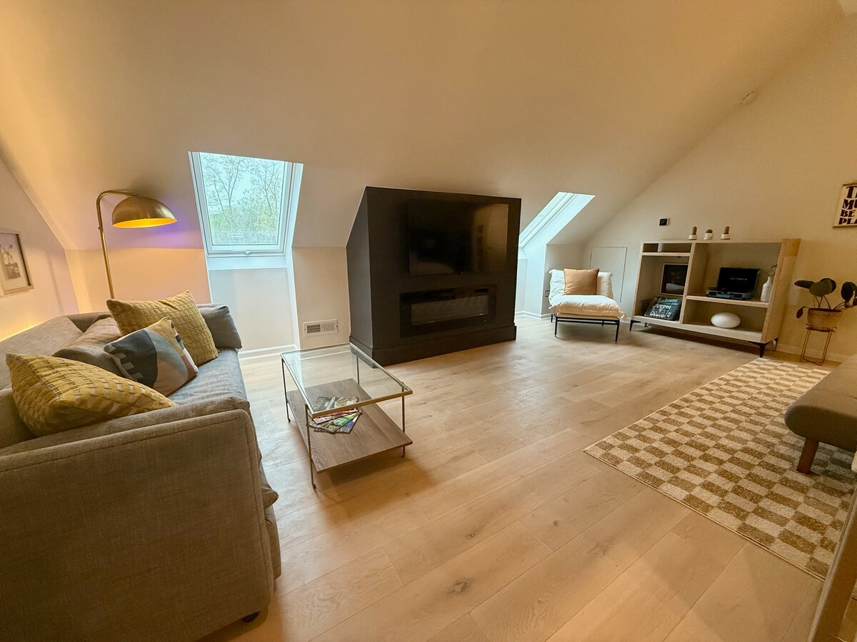 A bright living area features two skylights that enhance natural light. A cozy seating arrangement includes a gray sofa with colorful cushions and a glass coffee table. A modern fireplace serves as a focal point, complemented by a minimalist entertainment unit on one side.