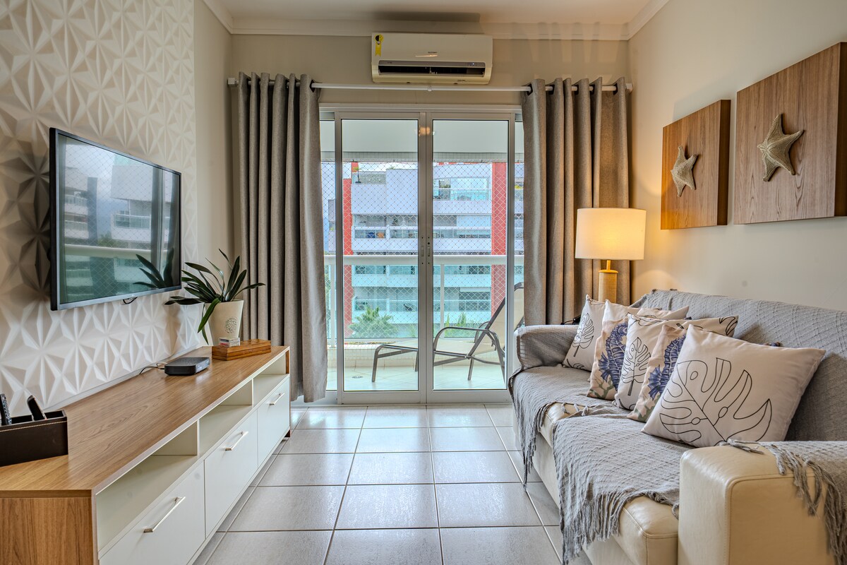 The living area features a comfortable sofa adorned with decorative pillows. A flat-screen TV is mounted on a wooden console. Natural light enters through large glass doors that lead to a balcony, framed by light curtains. Air conditioning is visible on the ceiling.