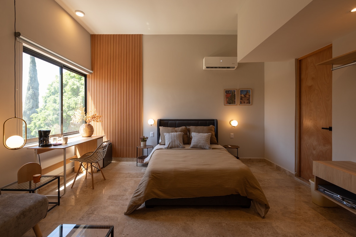 A well-lit room features a spacious bed with neutral bedding, complemented by decorative pillows. A desk area is positioned near the window, offering natural light. The walls are adorned with subtle decor, and a modern air conditioning unit is visible above the bed.