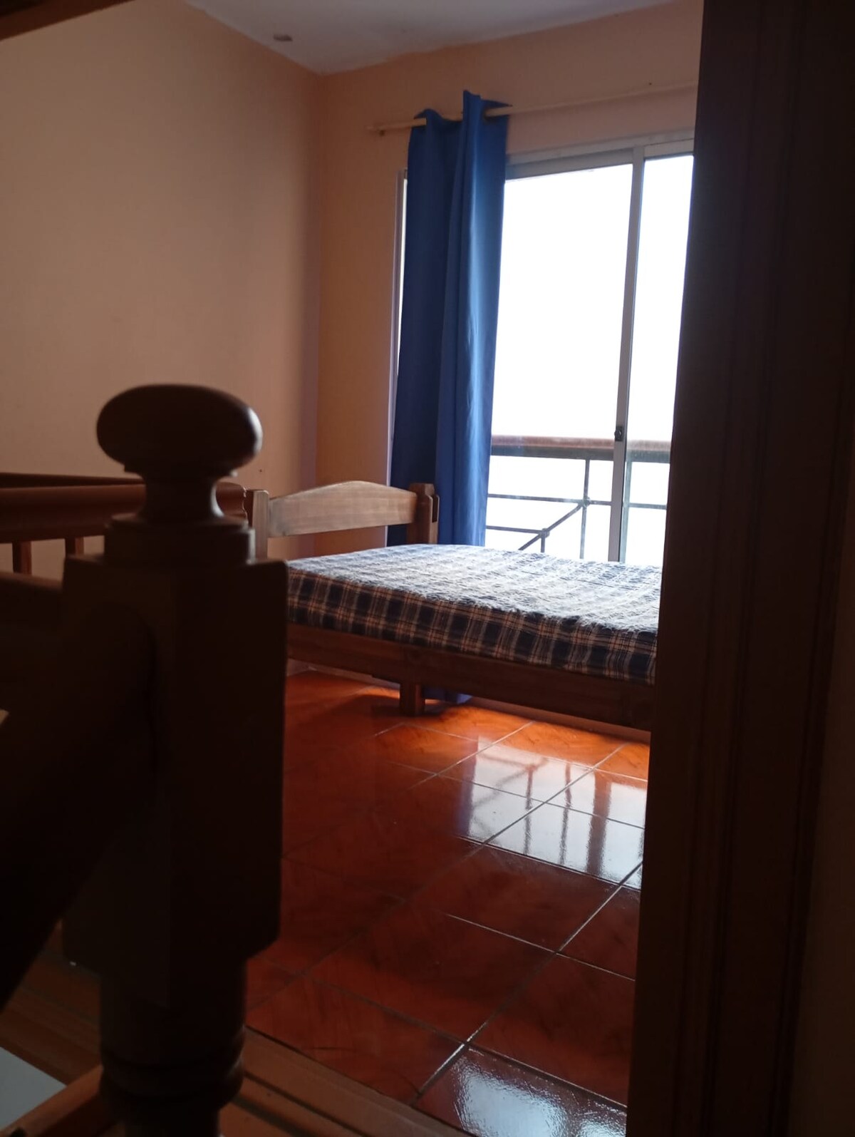 A simple bedroom is seen through a doorway, featuring a wooden bed with a checkered mattress. A bright blue curtain frames the window, allowing natural light to fill the space. The tiled floor reflects the light, enhancing the room's openness.