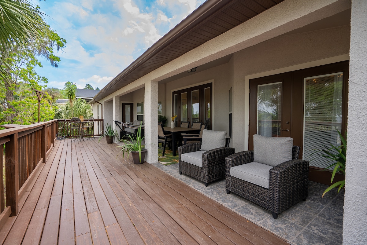 A spacious covered porch features comfortable wicker seating arrangements with soft cushions. Potted plants are positioned along the railing, enhancing the outdoorsy feel. The entrance opens to a bright interior, and the relaxing outdoor space offers a view of the surrounding trees.