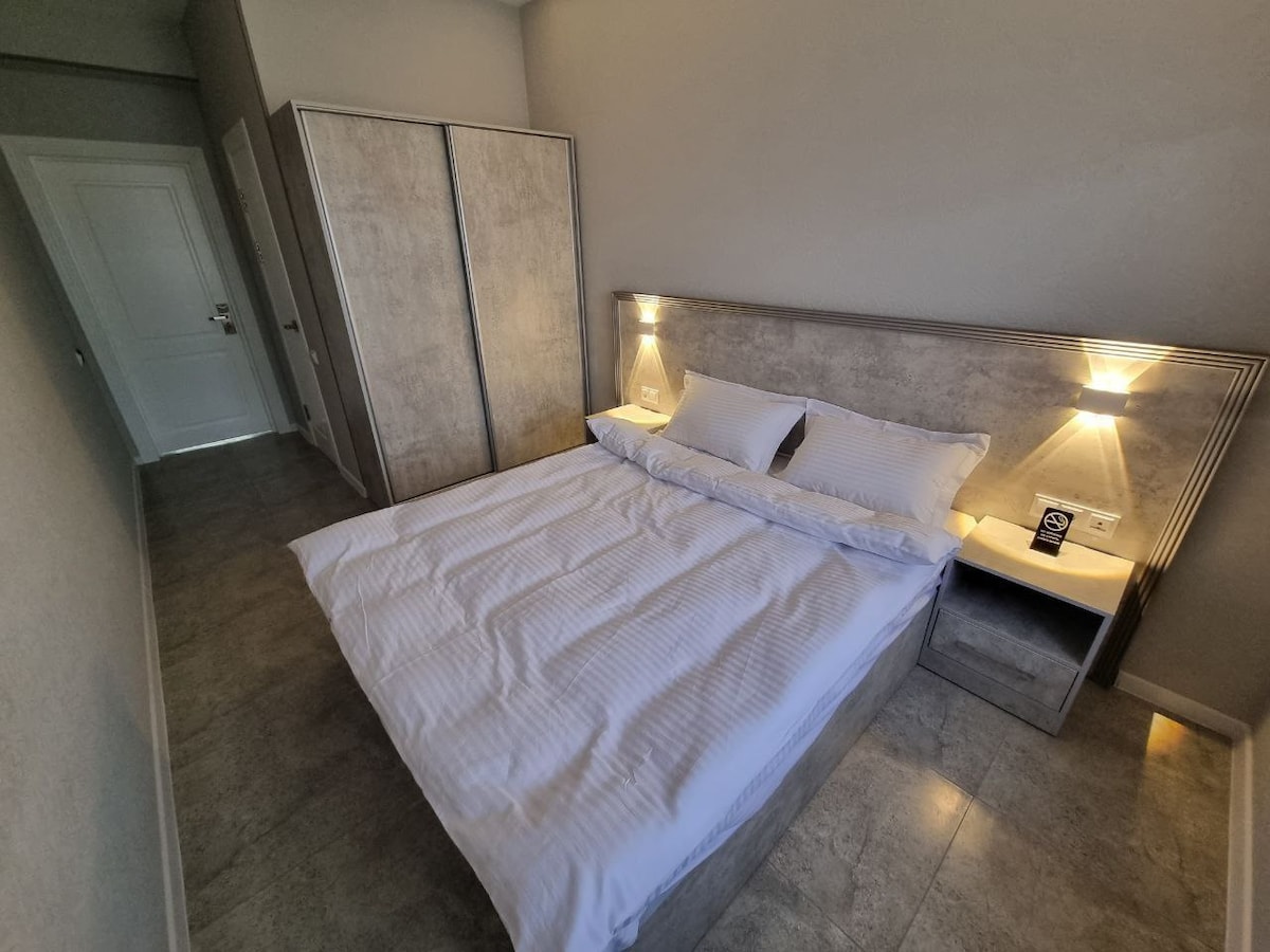 A modern bedroom is presented with a spacious bed featuring crisp white linens and subtle bedside lighting. A minimalist nightstand complements the bed, while an ample wardrobe in a neutral tone stands against the wall, enhancing the clean and organized aesthetic.