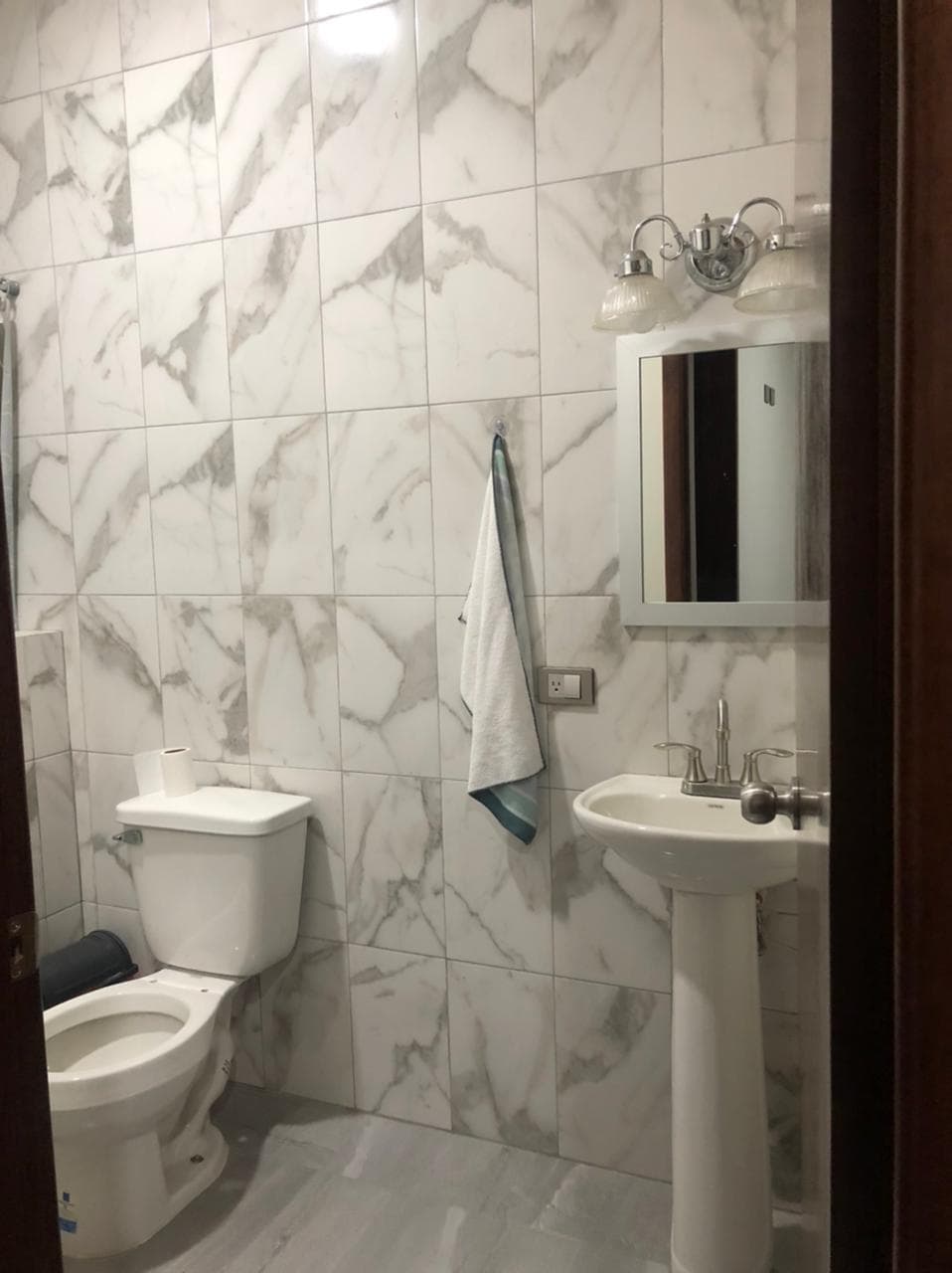 A bathroom is showcased with marble-patterned tiles covering the walls. A white toilet is visible beside a pedestal sink, which features a mirror above it. A hand towel hangs from a wall-mounted bar, while a bath mat lies on the floor.