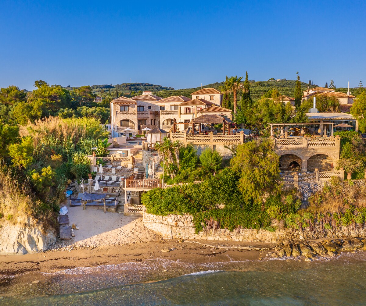 The aerial view captures the villa's spacious layout set against lush greenery. It features a beachfront area with sun loungers and a private path leading to the sandy shore. The structure blends traditional architecture with the natural surroundings, emphasizing tranquility and coastal living.