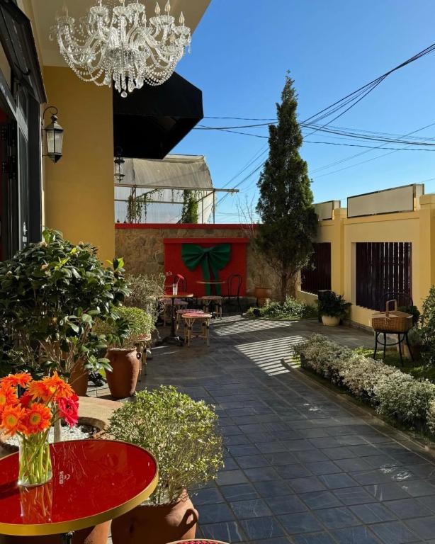 A charming outdoor area is presented with a vibrant red wall and decorative potted plants. A small table and chairs are placed on a tiled path, accompanied by a lush green garden featuring trim hedges and colorful flower arrangements, creating a serene ambiance.