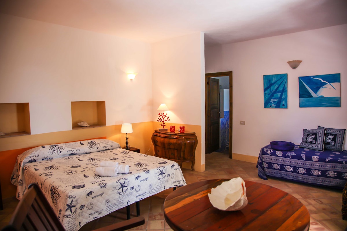 A spacious sleeping area is featured, with a bed covered in a white and blue patterned bedspread. A lamp provides soft illumination, and a wooden side table enhances the room's charm. Colorful wall art is displayed, and a cozy seating area is present in the corner.