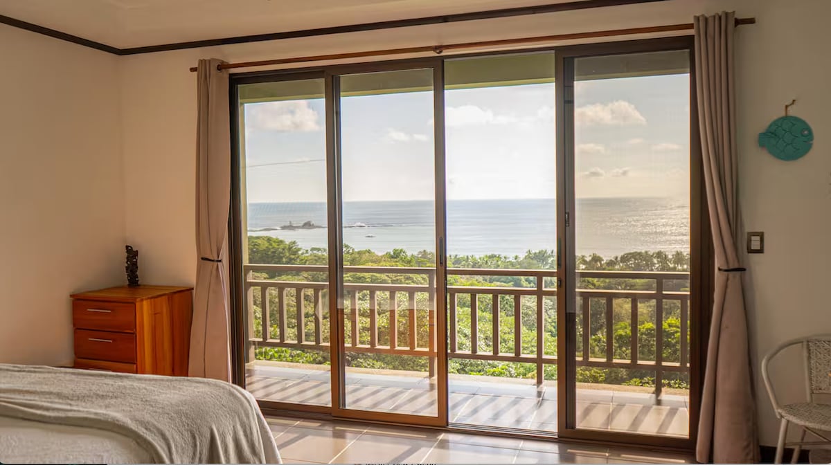A spacious bedroom features large sliding glass doors that open to a balcony with ocean views. Natural light floods the room, illuminating the wooden dresser and providing a tranquil backdrop of greenery and water. Soft curtains frame the view, enhancing the peaceful setting.